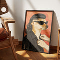 Business in the Front by Muse and Paw, a print of a dog in sunglasses, a black shirt and a mullet against a coral sky, framed and leaning against a wall.