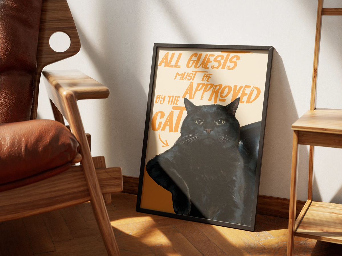 Door Policy by Muse and Paw, a print of a black cat with hand-lettered type reading all guests must be approved by the cat, framed and styled in a sunlit room with a leather armchair and wooden shelving.