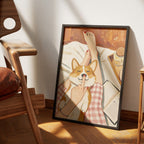 Five More Minutes by Muse and Paw, an overhead print of a corgi in bed with white sheets, pink gingham pyjamas and a finger on his nose, framed and leaning against a wall.