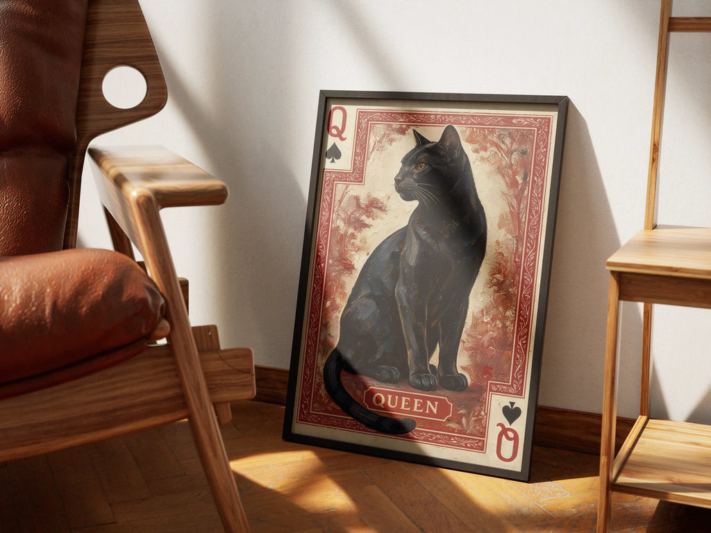 Her Majesty by Muse and Paw, a print of a black cat as the Queen of Spades on a red card, framed and styled in a sunlit room with a leather armchair and wooden shelving.