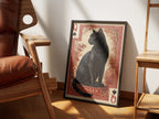 Her Majesty by Muse and Paw, a print of a black cat as the Queen of Spades on a red card, framed and styled in a sunlit room with a leather armchair and wooden shelving.