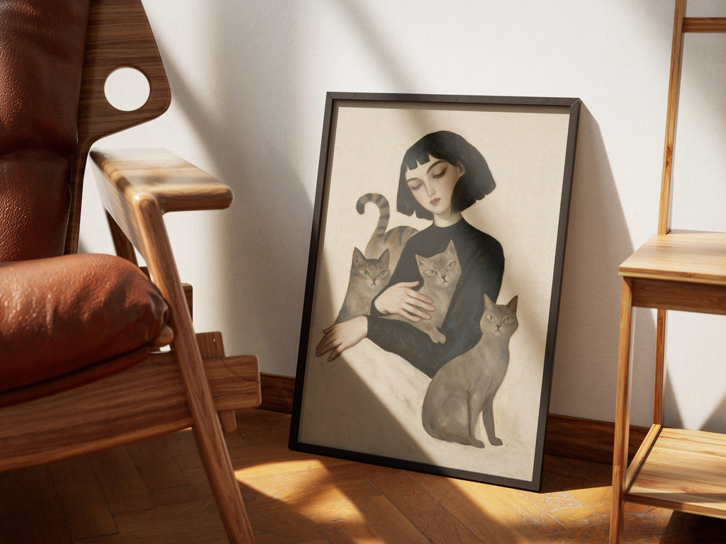 Menagerie by Muse and Paw, a portrait print of a woman holding a tabby, a grey tabby, and a smoke-grey cat, styled in a sunlit room with a leather armchair and wooden chairs.
