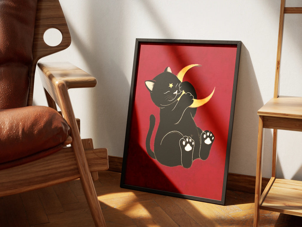 Moonstruck by Muse and Paw, an illustrated print of a black cat holding a gold crescent moon on a deep red ground, framed and styled in a sunlit room with a leather armchair and wooden chairs.