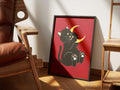 Moonstruck by Muse and Paw, an illustrated print of a black cat holding a gold crescent moon on a deep red ground, framed and styled in a sunlit room with a leather armchair and wooden chairs.