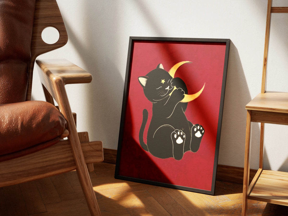 Moonstruck by Muse and Paw, an illustrated print of a black cat holding a gold crescent moon on a deep red ground, framed and styled in a sunlit room with a leather armchair and wooden chairs.