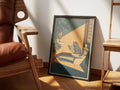 Occupied by Muse and Paw, a print of a grey cat reading the newspaper on the toilet in a sunlit bathroom, framed and leaning against a sunlit wall beside a leather chair and wooden shelving.