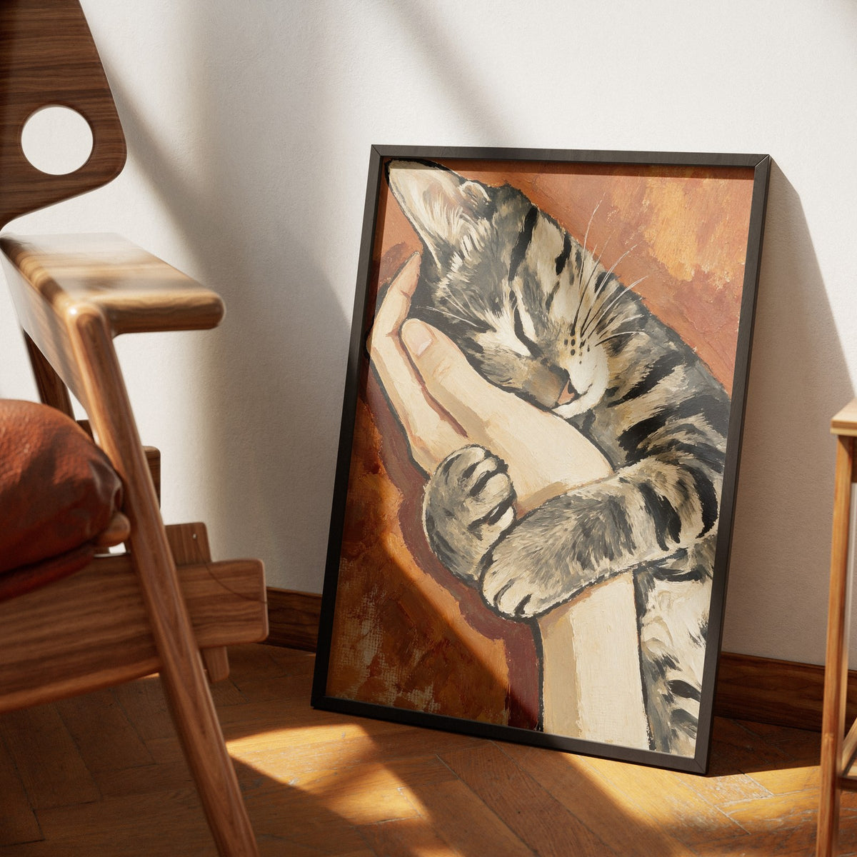 Safe by Muse and Paw, a painterly print of a tabby cat asleep in an open hand against a warm rust background, framed and leaning against a wall.