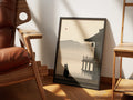 Still Water by Muse and Paw, a print of a black cat on a wooden jetty looking out over a misty lake beside a Japanese temple, framed and leaning against a sunlit wall beside a leather chair and wooden shelving.