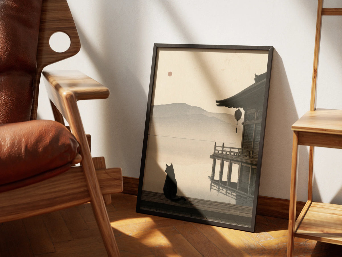 Still Water by Muse and Paw, a print of a black cat on a wooden jetty looking out over a misty lake beside a Japanese temple, framed and leaning against a sunlit wall beside a leather chair and wooden shelving.