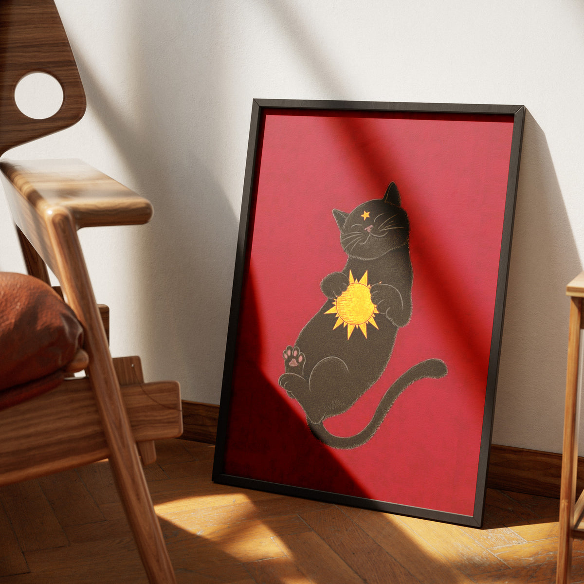 Sunspot by Muse and Paw, a print of a black cat lying on its back on a deep red ground, holding a small golden sun against its chest, framed and leaning against a wall.
