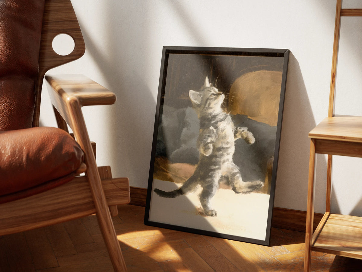Two Step by Muse and Paw, a print of a silver tabby kitten dancing on its back legs, framed and styled in a sunlit room with a leather armchair and wooden shelving.