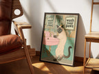 Wash Your Paws by Muse and Paw, a print of a Siamese cat washing her paws at a pink basin under a wash your paws sign, framed and leaning against a sunlit wall beside a leather chair and wooden shelving.